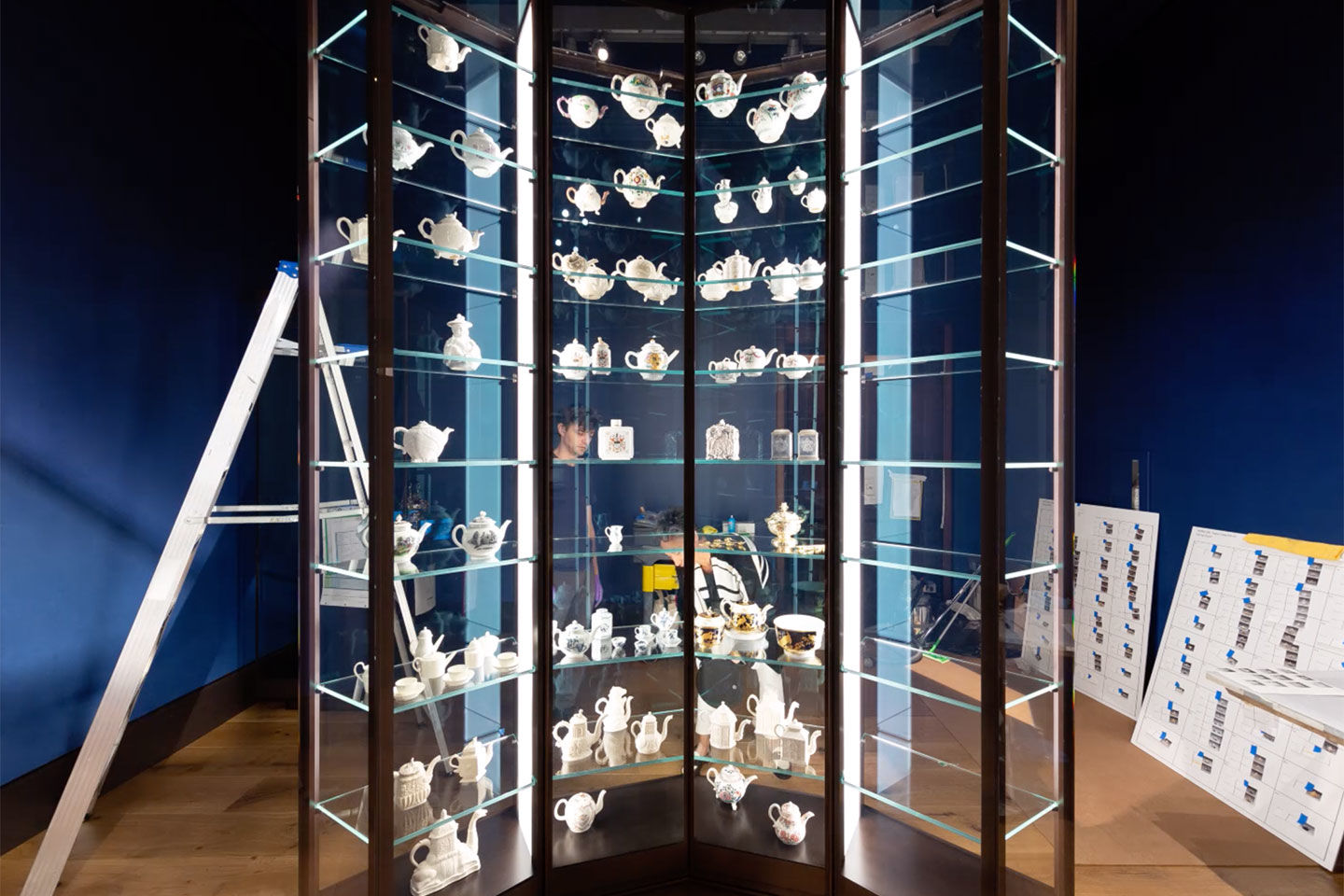 Semi-circular, glass display case in the process of being filled with many different teapots.